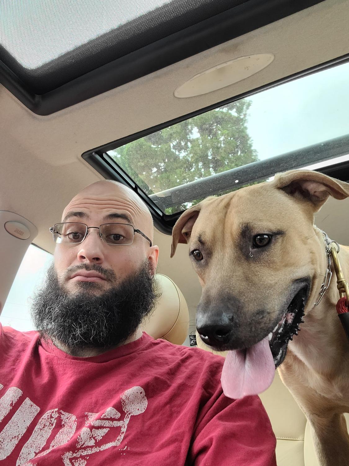 Jeremy Ryan in a car with a tan dog leaning into the frame, tongue out.